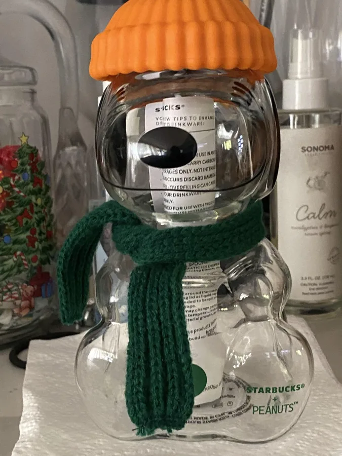 A Snoopy-shaped glass cup with an orange beanie, green knitted scarf and 'STARBUCKS® PEANUTS' printed on the belly.