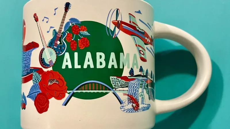 The Alabama Discovery Series mug features images of a guitar, airplane, football helmet, beachgoers, and a rocket launch scene, representing the state's culture and attractions