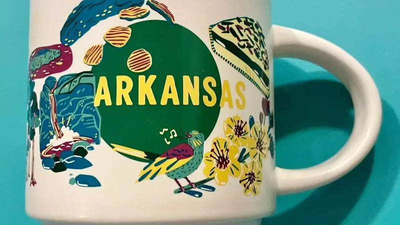 A Starbucks Arkansas Discovery Series mug featuring the state’s Capitol building, a violinist, flowers, diamonds, and a colorful bird, representing Arkansas’s culture and nature.