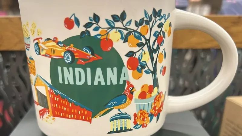 The Indiana Discovery Series mug features a red race car, peach tree, bird, classical building, and a woman sewing alongside books and a quilt.