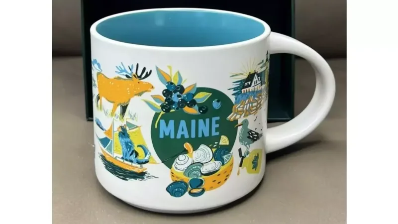 The Maine Discovery Series mug features a colorful design including a moose, blueberries, sailboats, lighthouses, and a basket of clams, celebrating the state's coastal beauty and nature.