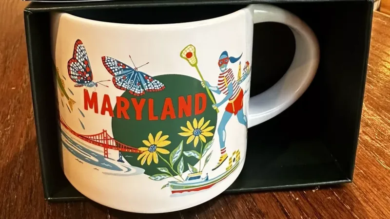 Maryland-themed mug featuring a lacrosse player, blue crab, state bird, and Annapolis State House, with the Chesapeake Bay Bridge in the background