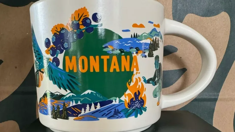 A Starbucks mug featuring Montana's scenic design with mountains, a bear, an eagle carrying books, blueberries, and a camper by a campfire.