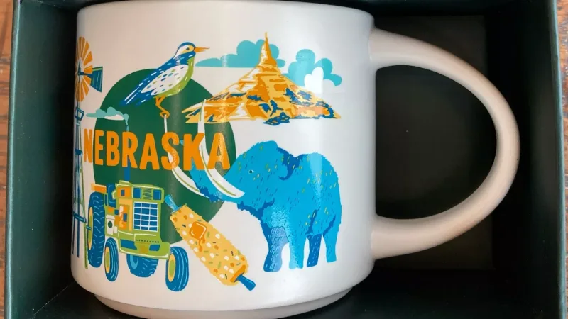 A Nebraska-themed Starbucks mug featuring a woolly mammoth, corn-on-the-cob, a tractor, windmill, frying pan, and scenic landscapes in vibrant colors.