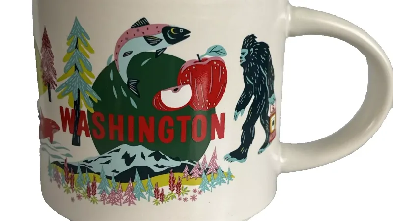 A white mug with colorful illustrations of Washington state landmarks, including Mount Rainier, an orca, a red apple, Bigfoot, and outdoor adventure scenes.